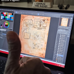 Hand holding up a thumbs-up in front of a Dell 2-in-1 laptop running Clip Studio Paint. The screen shows a detailed fantasy map of a cultist base. A Garfield 'Compute This, Sucker' figurine sits on top of the laptop in a cozy setup.